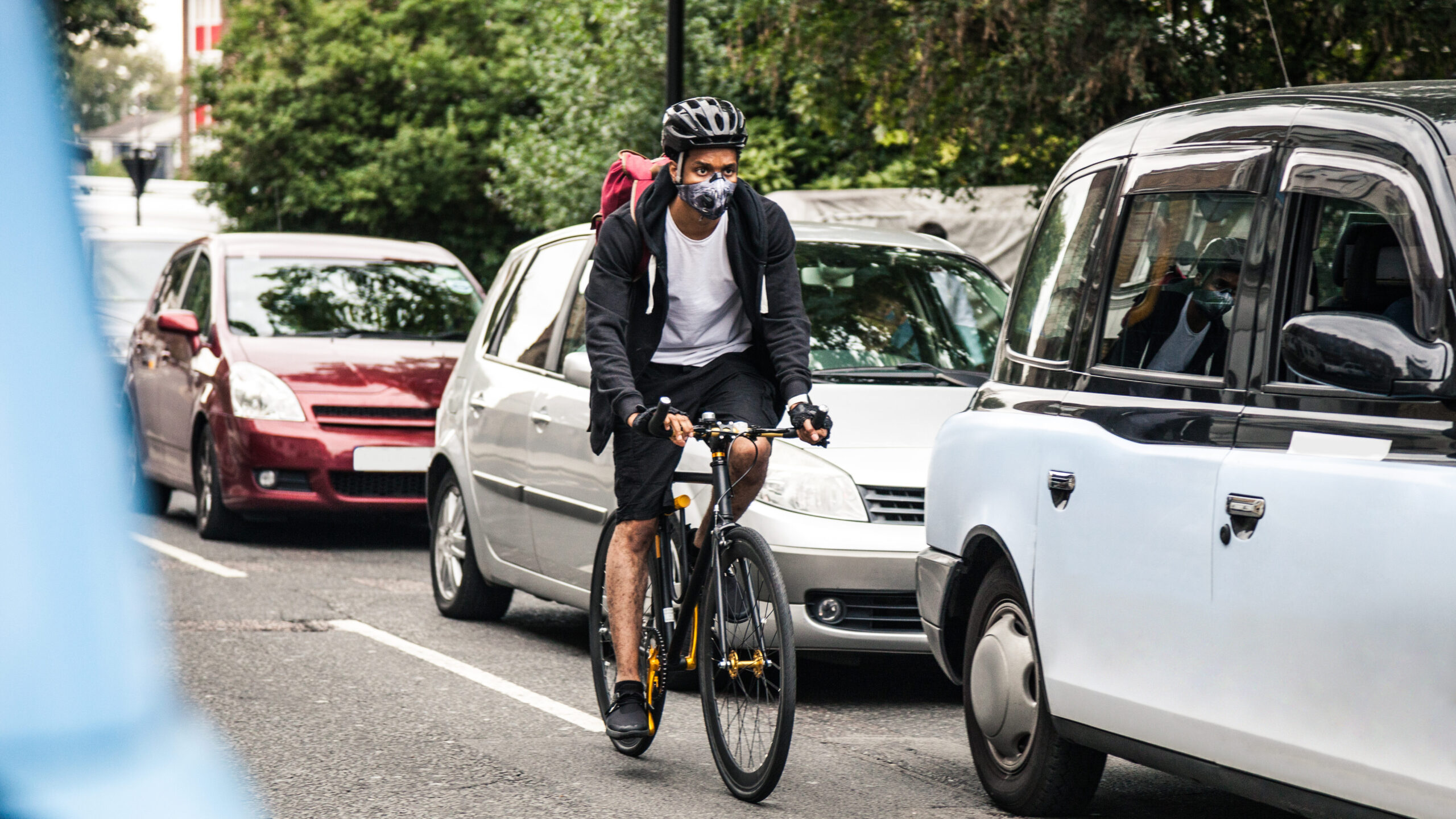A man cycling on a road congested with cars. The man is wearing a lower face mask for better air ventilation.